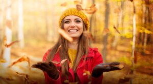 Strahlende junge Frau mit gelber Mütze und rotem Mantel wirft Herbstlaub in die Luft und lacht im herbstlichen Wald.